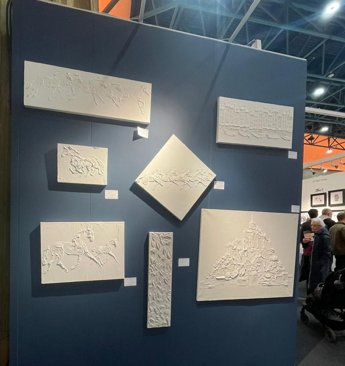 White textured art pieces on a blue wall at an art fair. People viewing art in background.