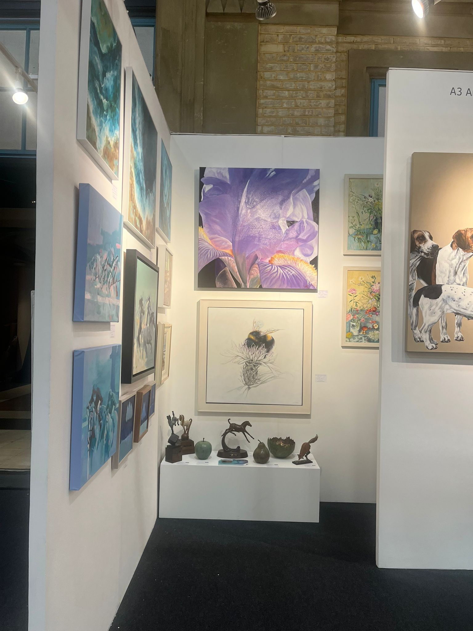 Art gallery booth with paintings and sculptures on display. White walls, dark carpet, various art styles.