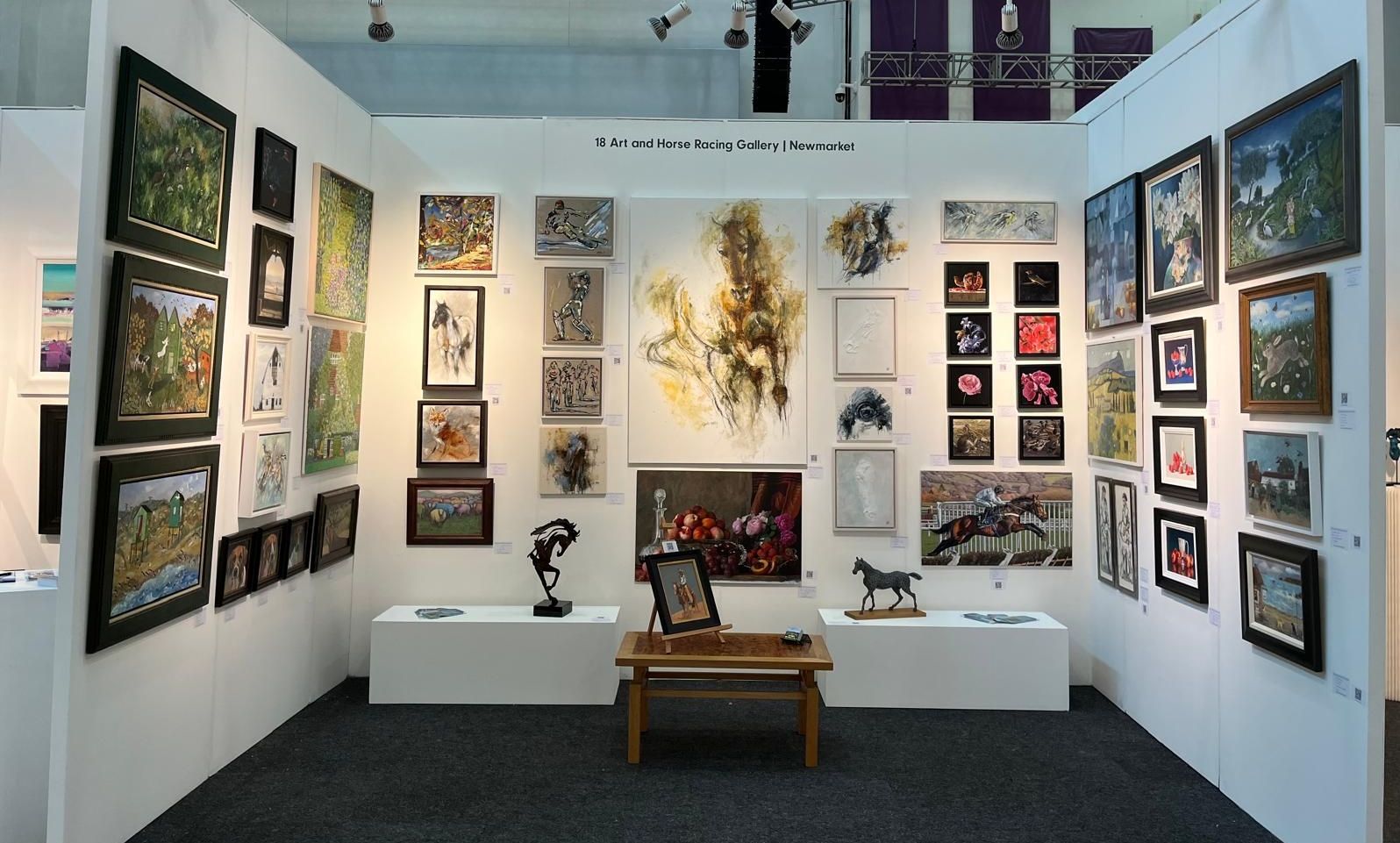 Art gallery booth with framed artwork on white walls, sculptures, and a small table.