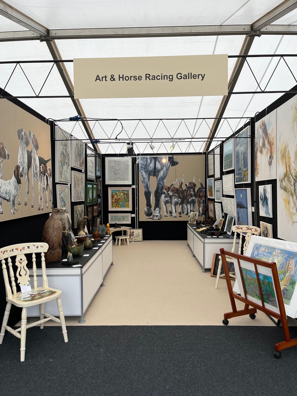 Art gallery booth: Horse racing art displayed, black & white photos, paintings. Beige interior, white tables, black carpet.