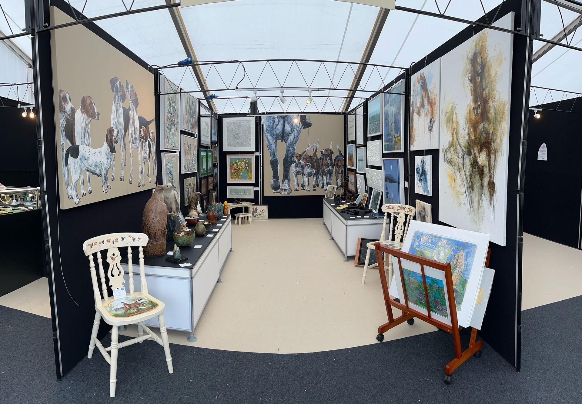 Art fair booth displaying paintings, pottery, and prints, with a white chair, on display under a tent.