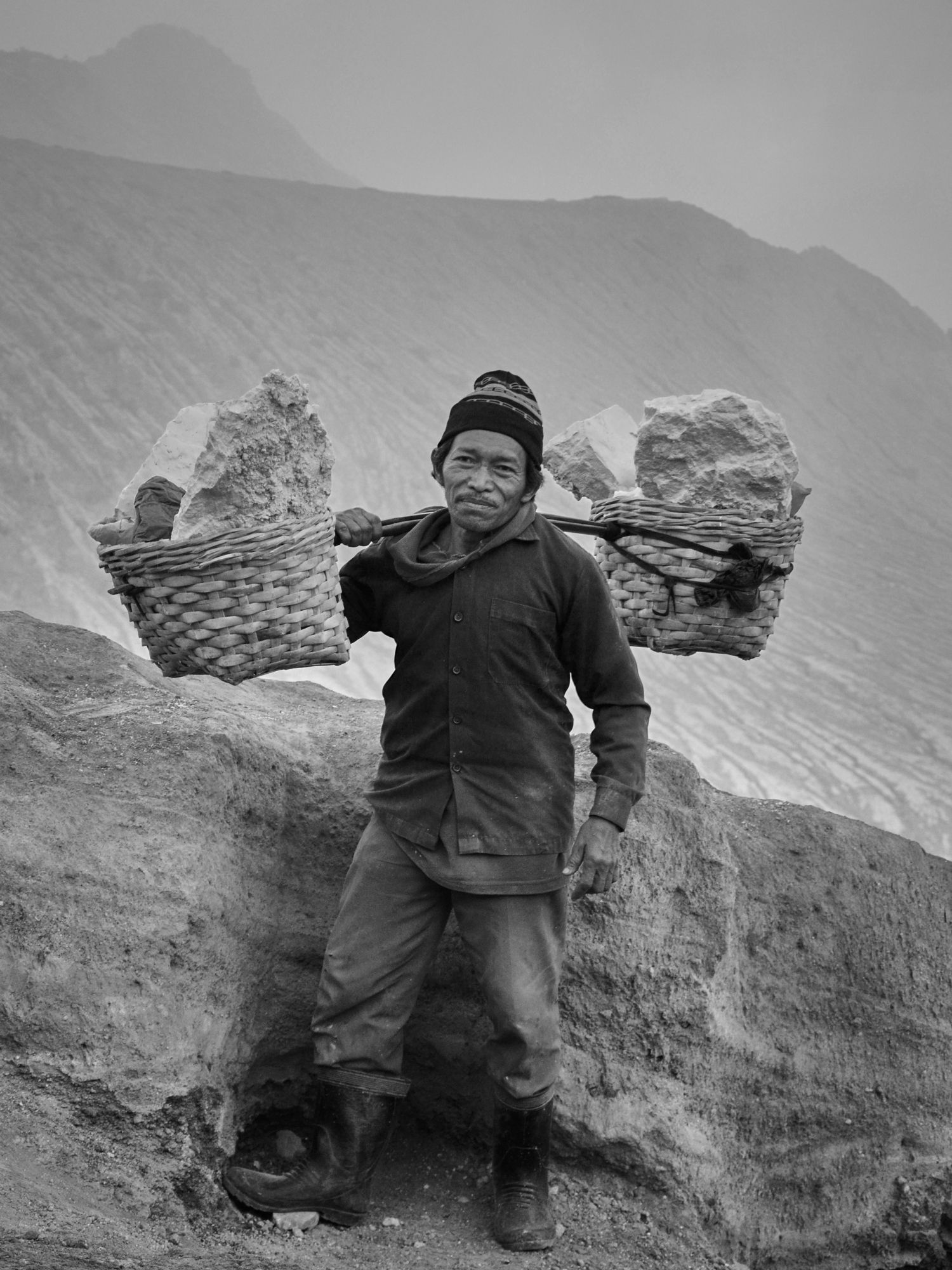 Man in warm clothing carries two baskets of rocks across his shoulders, standing on a rocky landscape with a mountain in the background.