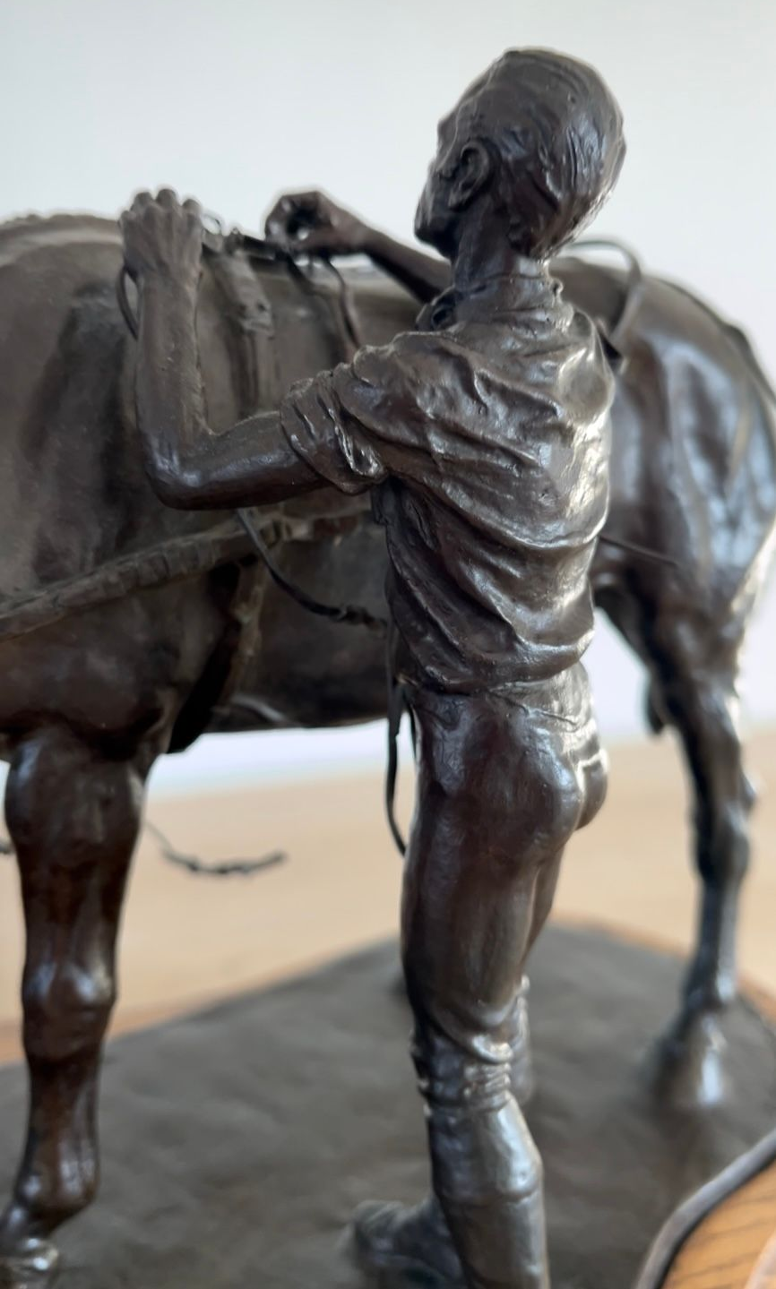 Bronze sculpture of a person adjusting a saddle on a horse; brown tones, indoor setting.