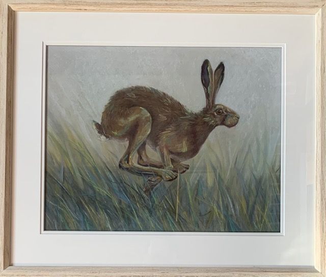 Brown hare leaping through tall green and blue grass, set in a light-colored, framed painting.