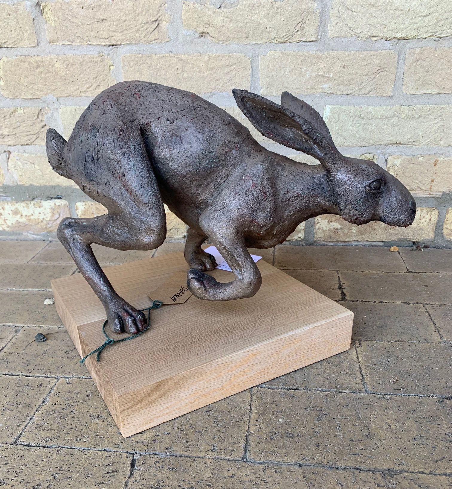 Bronze hare sculpture leaping from a wooden block, against a brick wall.