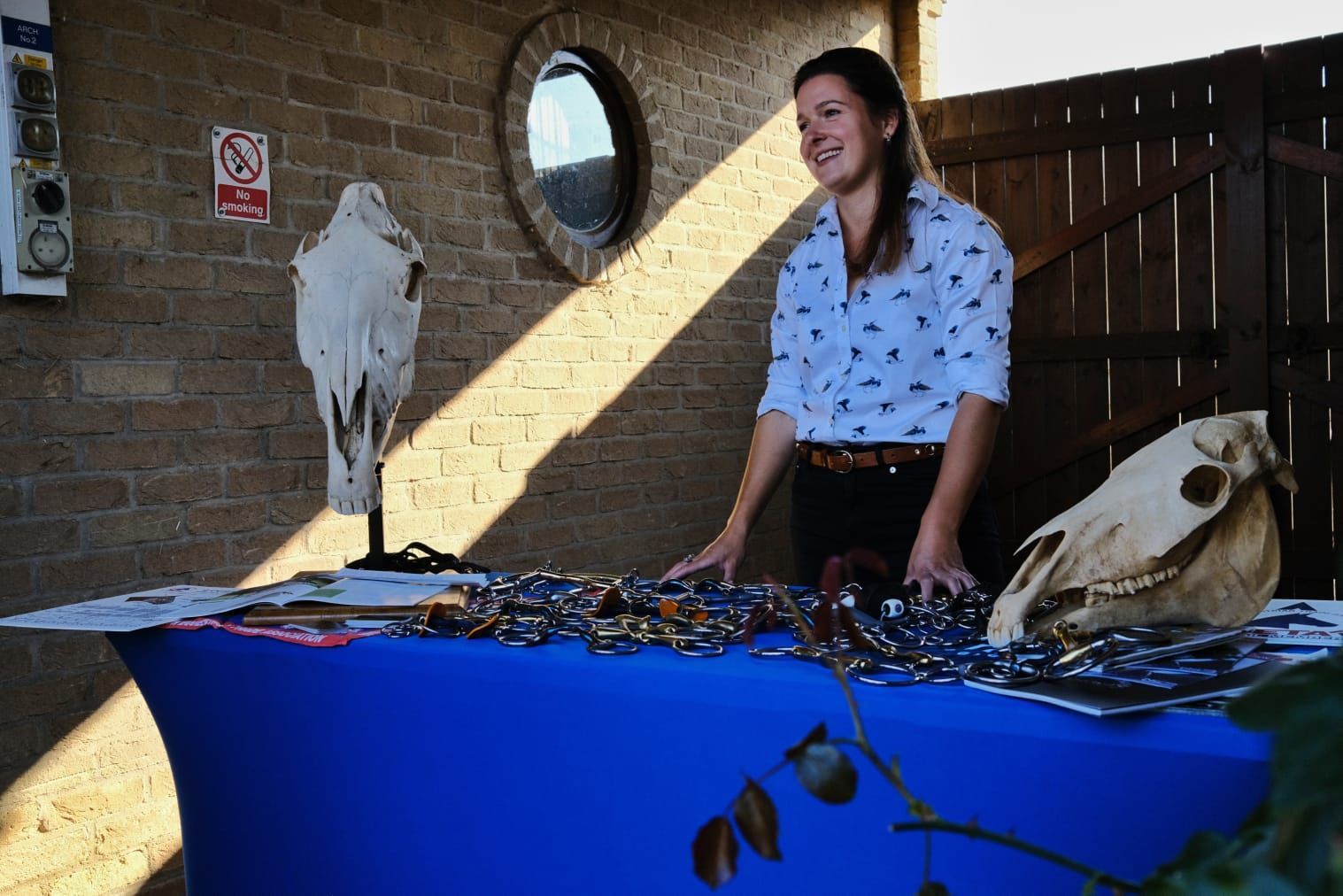 A woman is standing in front of a table with horse head skulls and bits on it.