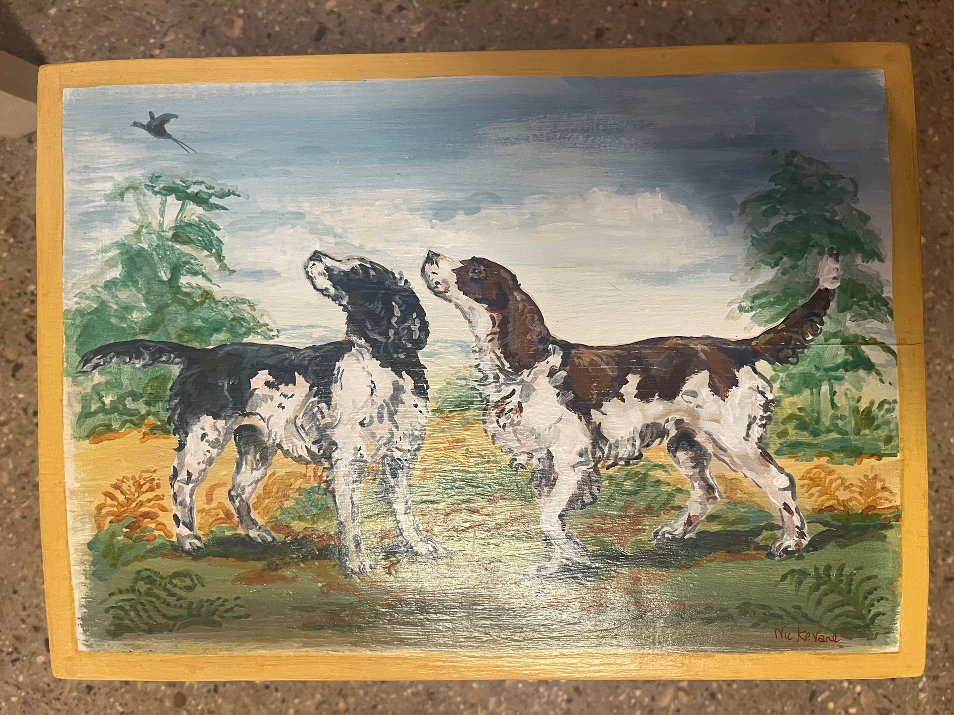 Two dogs looking up at a bird in a field; green, brown and white dogs with trees, a blue sky and a yellow frame.