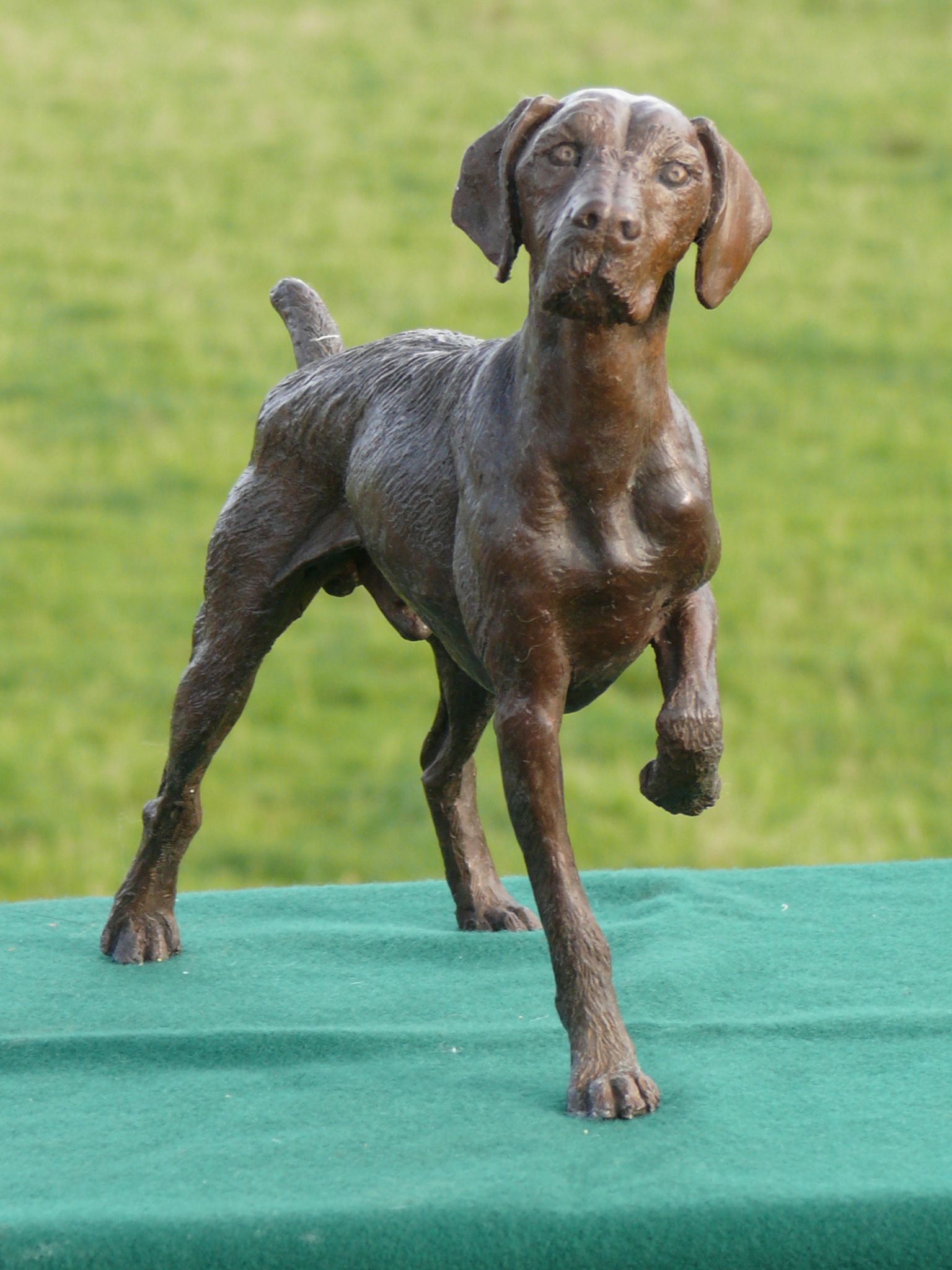 Bronze sculpture of a dog, standing on a wooden base, in front of a brick wall.