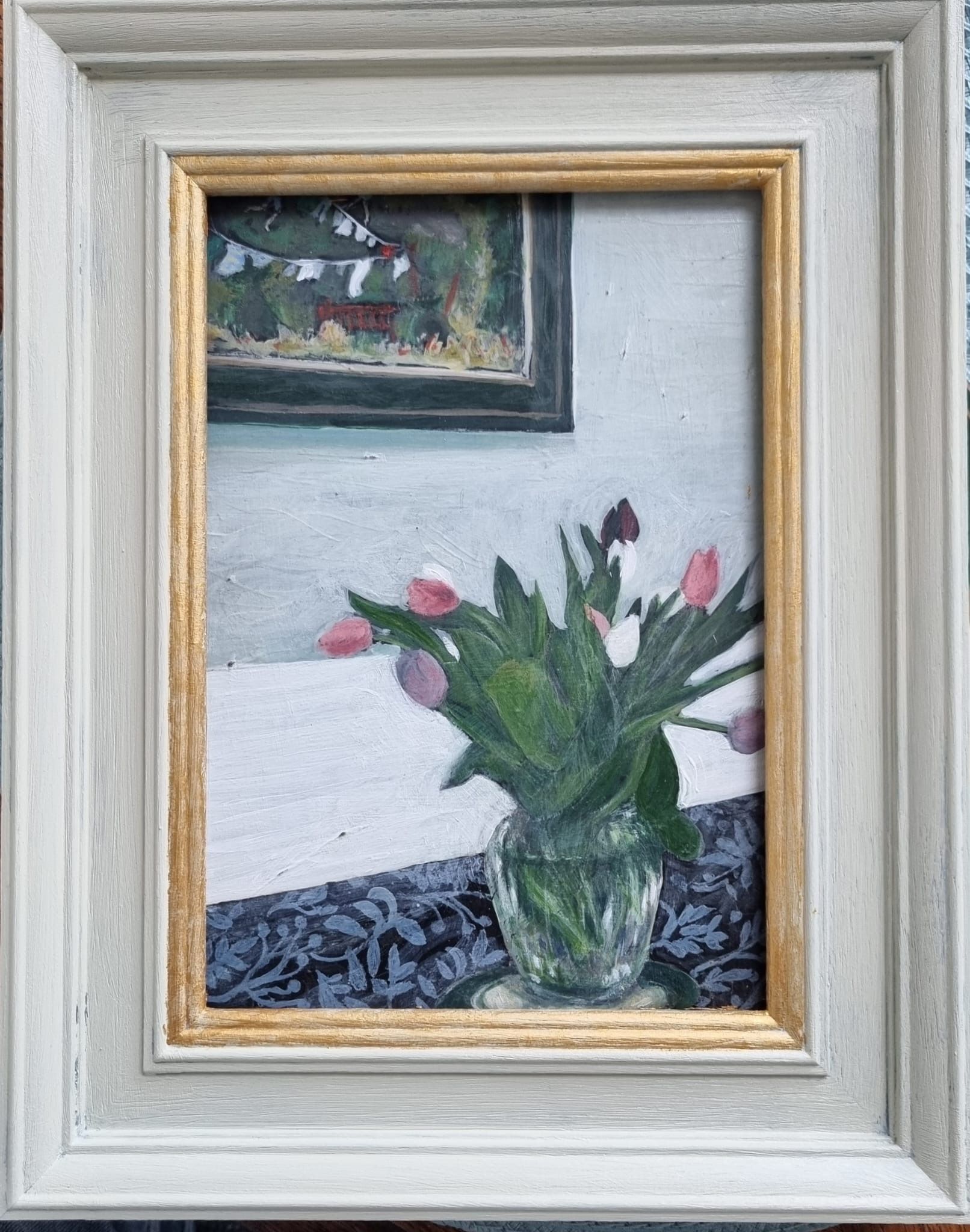 Painting of tulips in a glass vase on a patterned table, with a framed artwork above.
