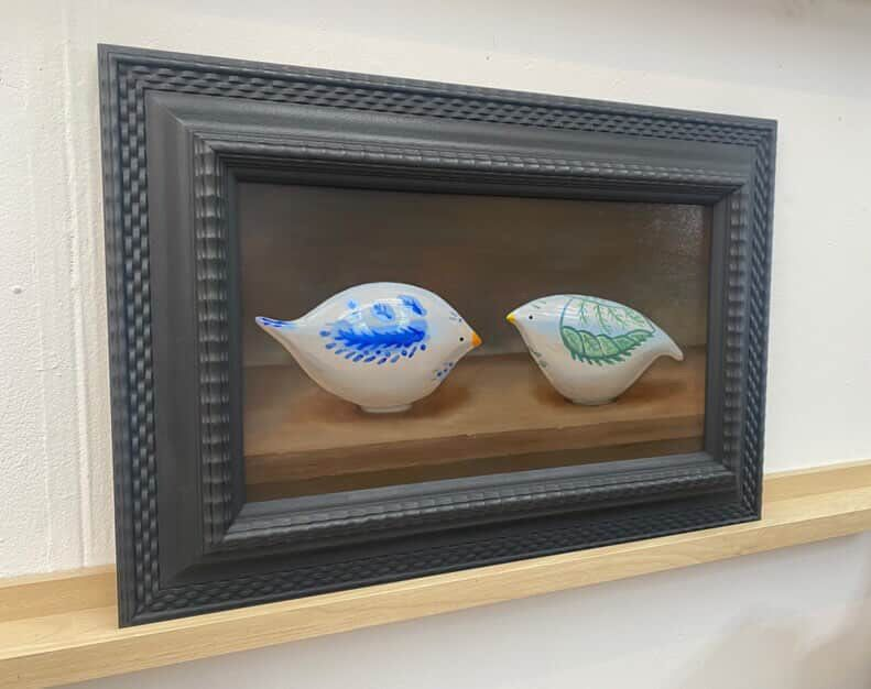 Two decorated white shell ornaments in a black frame, one blue and one green, on a brown background