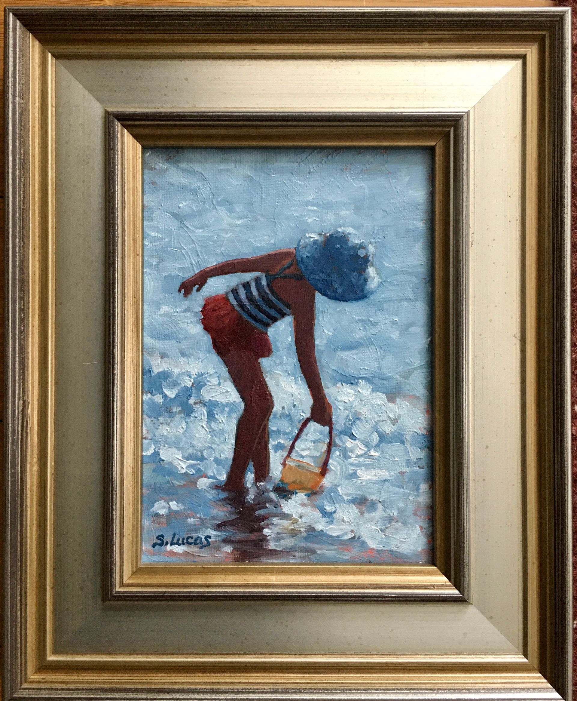 A painting in a silver frame shows a person in a striped top and red bottoms holding a bucket while wading in the ocean.