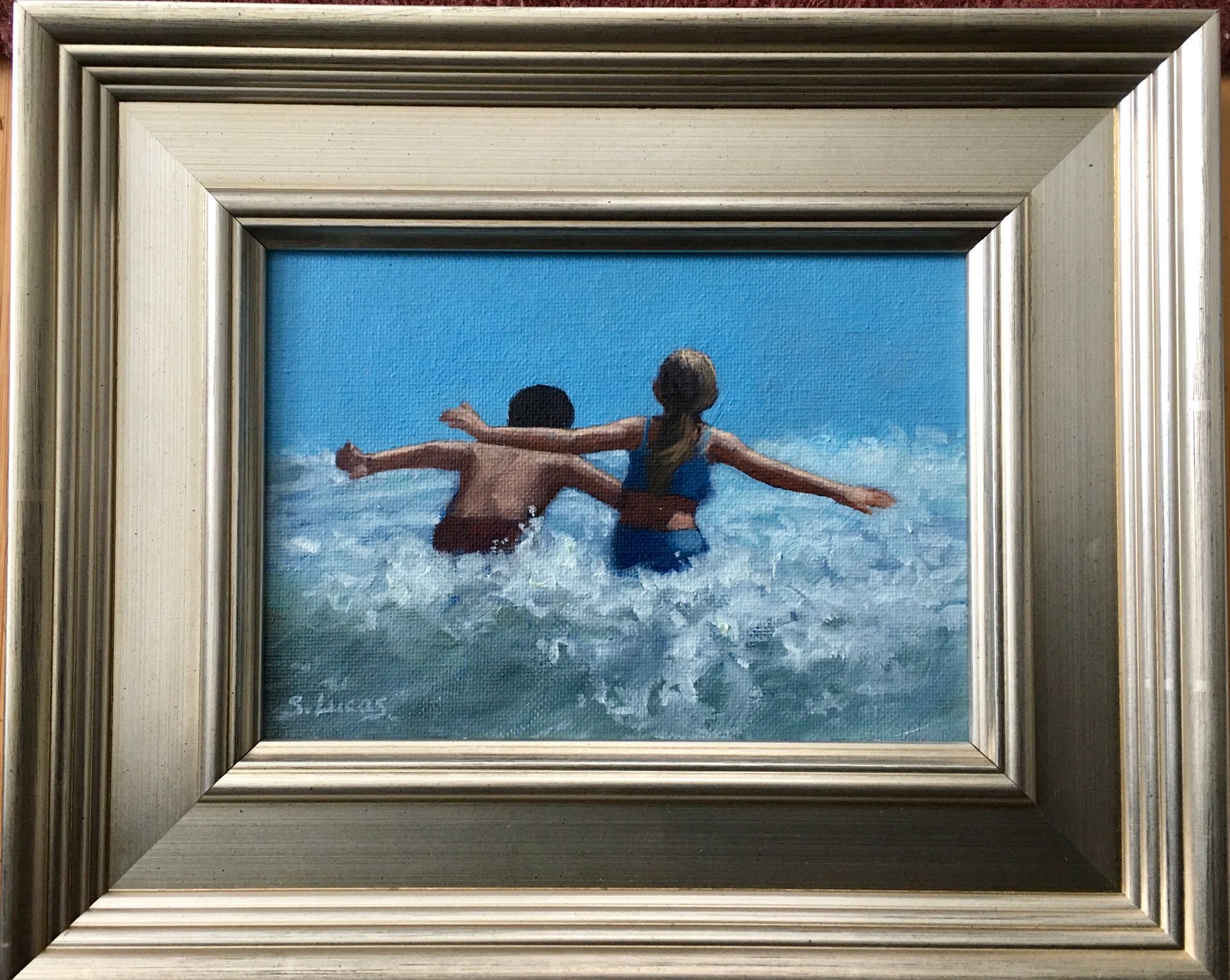 A framed oil painting, two people stretch out their arms while wading into ocean waves under a bright blue sky.