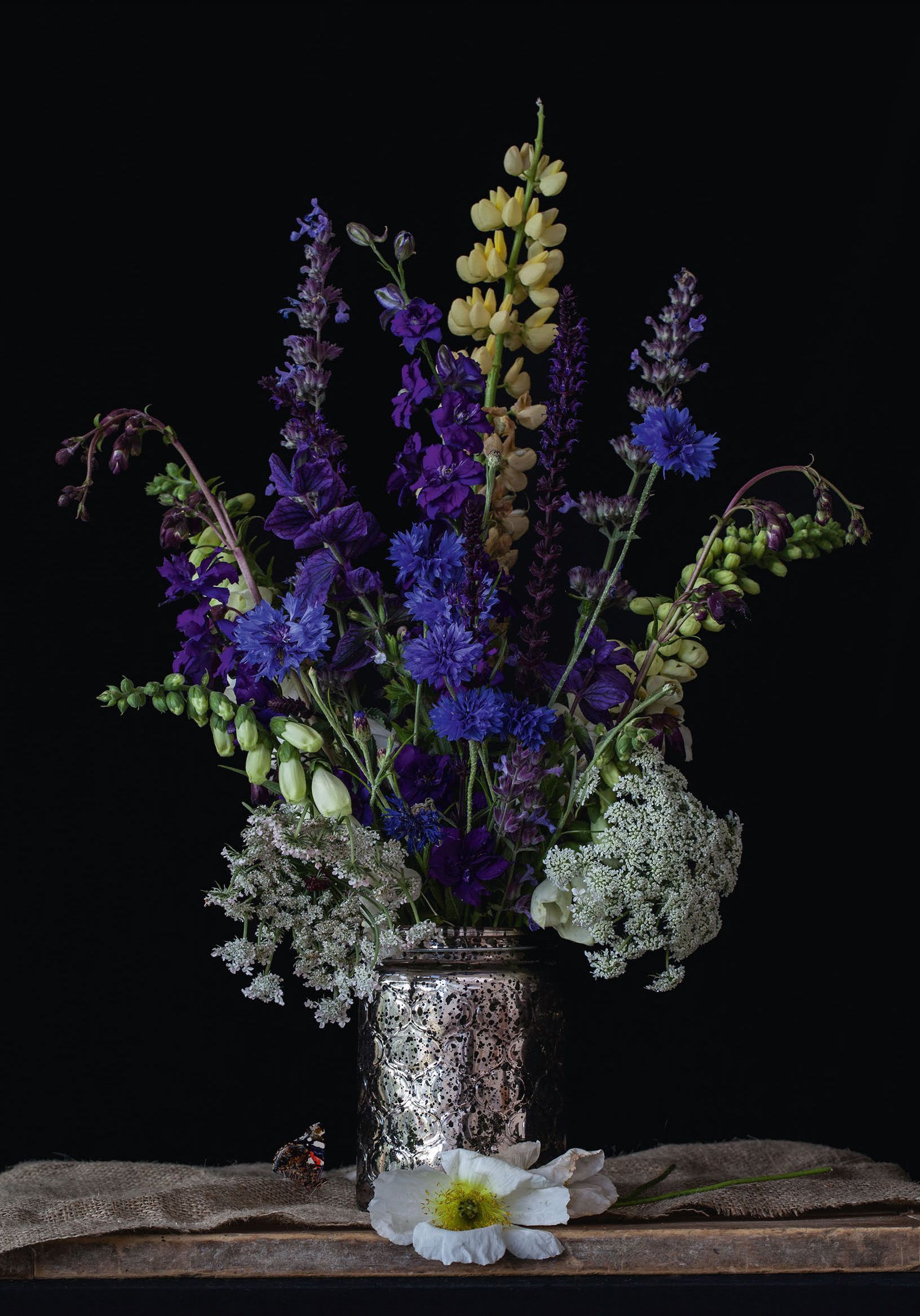 Bouquet of blue, purple, and yellow flowers in a silver vase, on a wooden surface against a black backdrop.