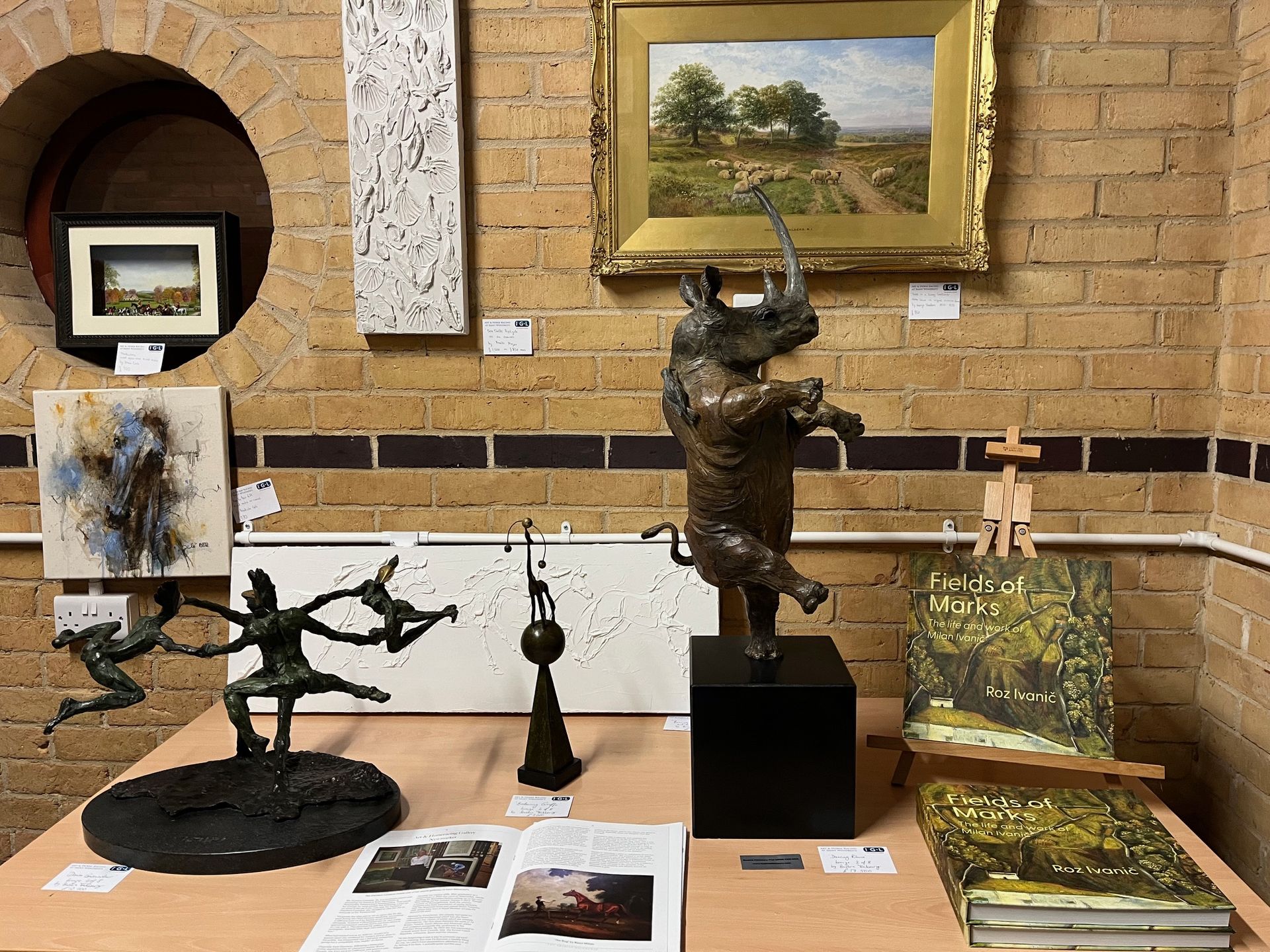 Art display in a room with brick wall. Sculptures of figures and rhino, paintings, and drawings on display.