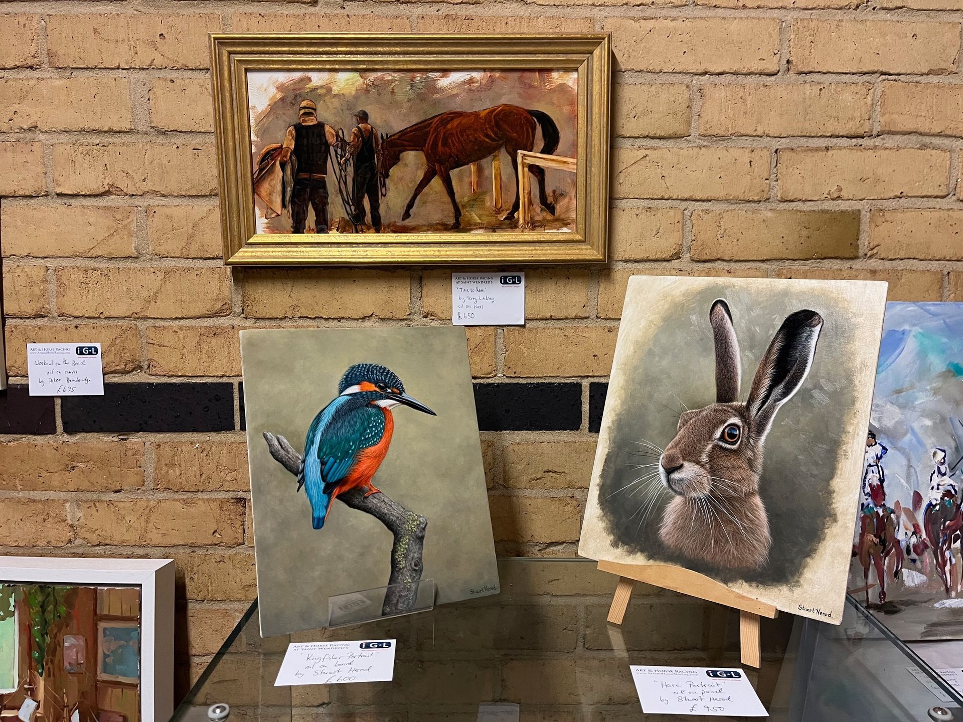 Artwork display: horse painting, kingfisher on a branch, hare on easel, brick wall background.