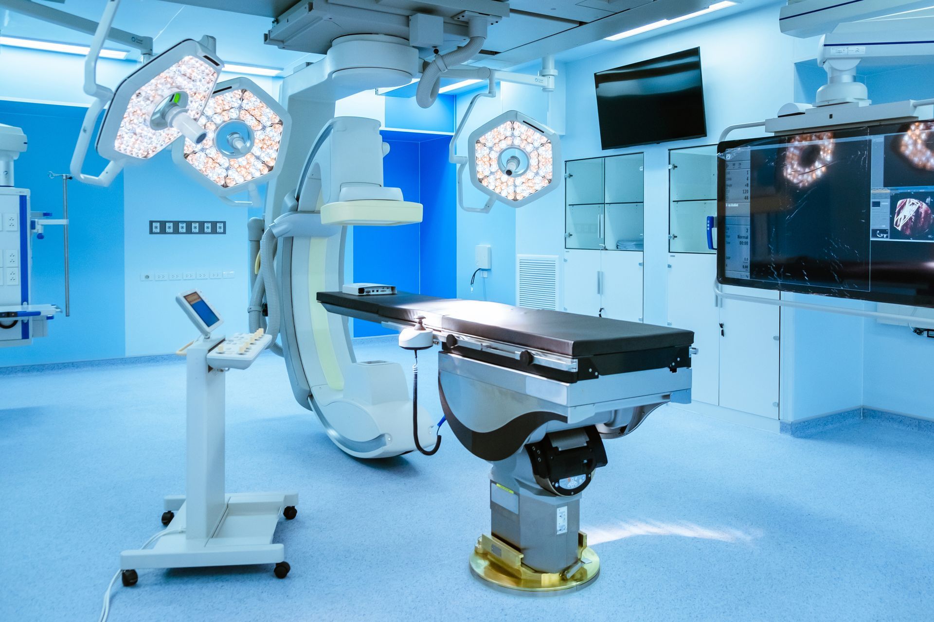 Operating room with surgical lights, equipment, and a table, in blue and white tones.