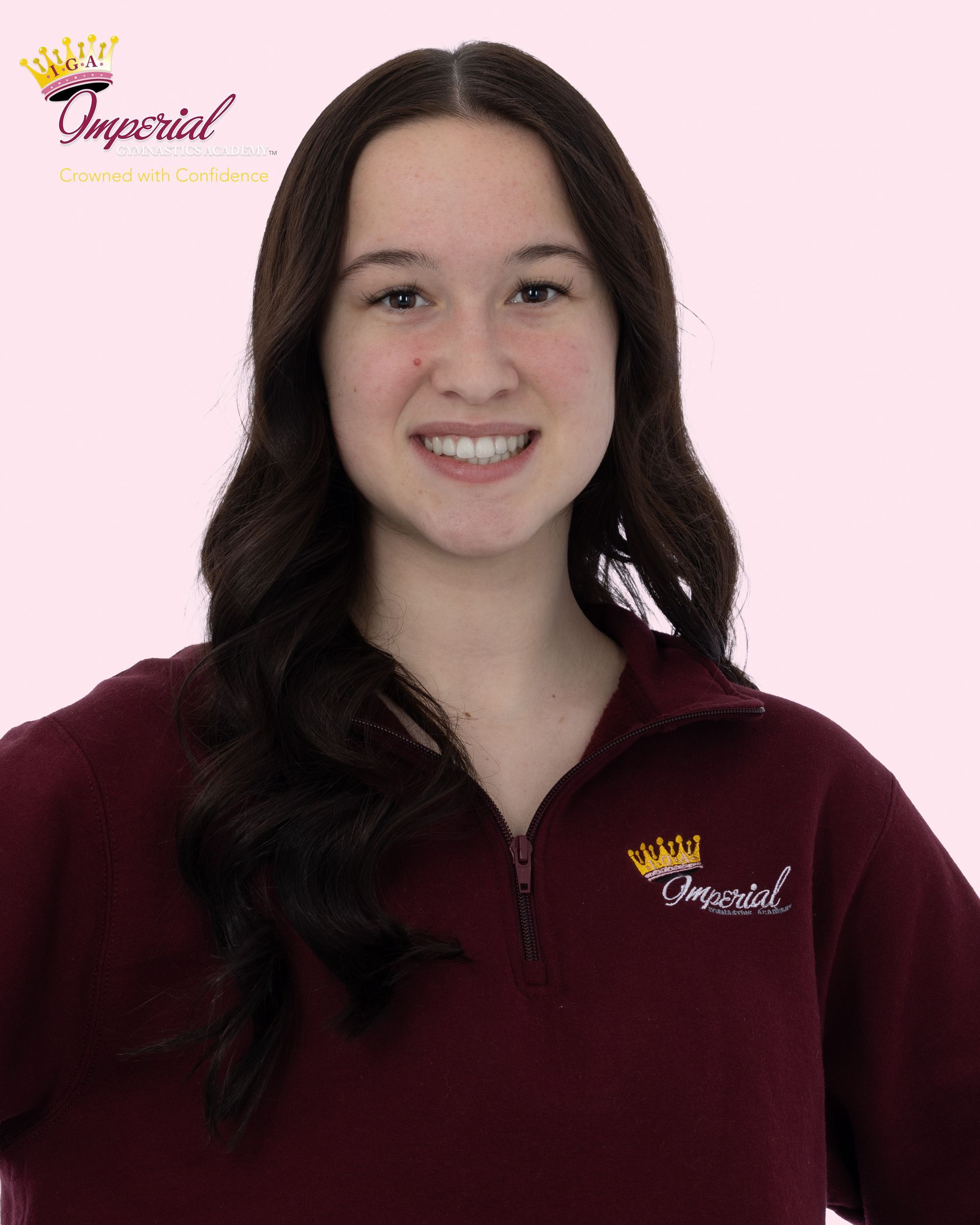 Woman with long dark hair smiles, wearing a pink t-shirt with text, necklace, against a white backdrop.