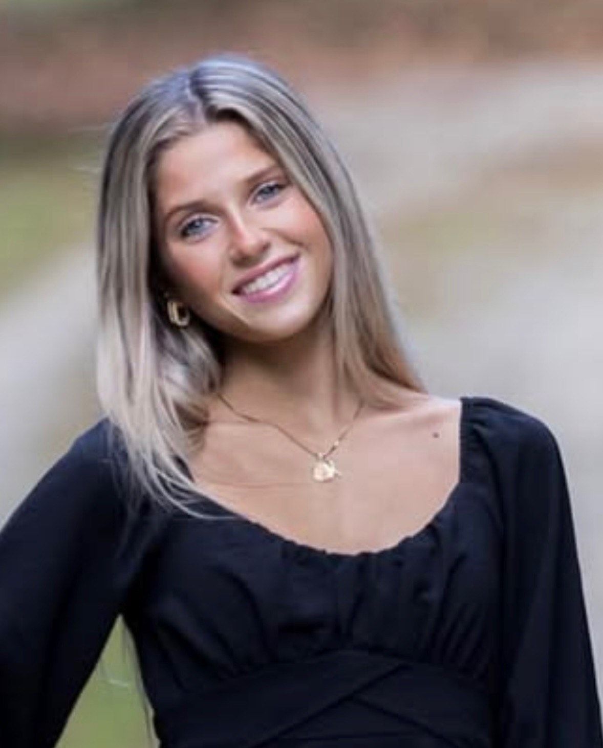 Blonde woman smiling, wearing a black top and a gold necklace, outdoors.
