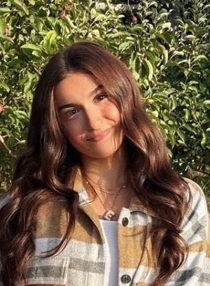 Woman with long, wavy brown hair smiles at the camera, wearing a plaid shirt and necklaces; outdoors.