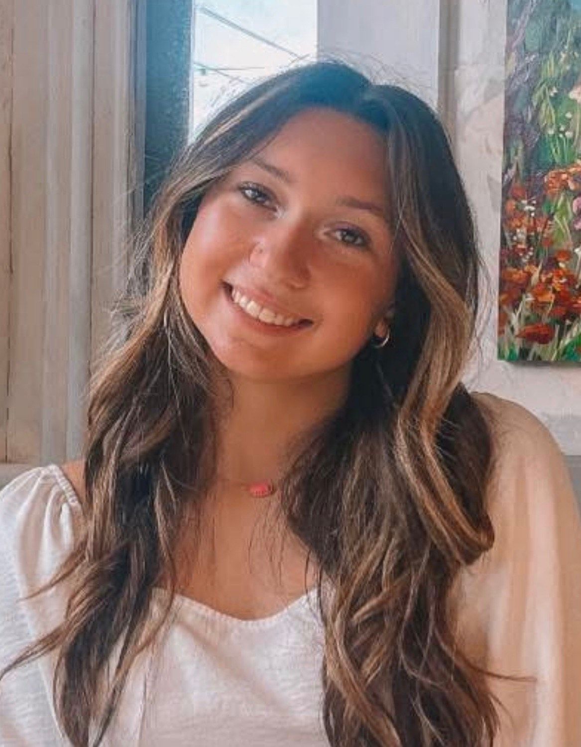 Woman with long brown hair smiling at the camera, wearing a white shirt, pink necklace, seated indoors.