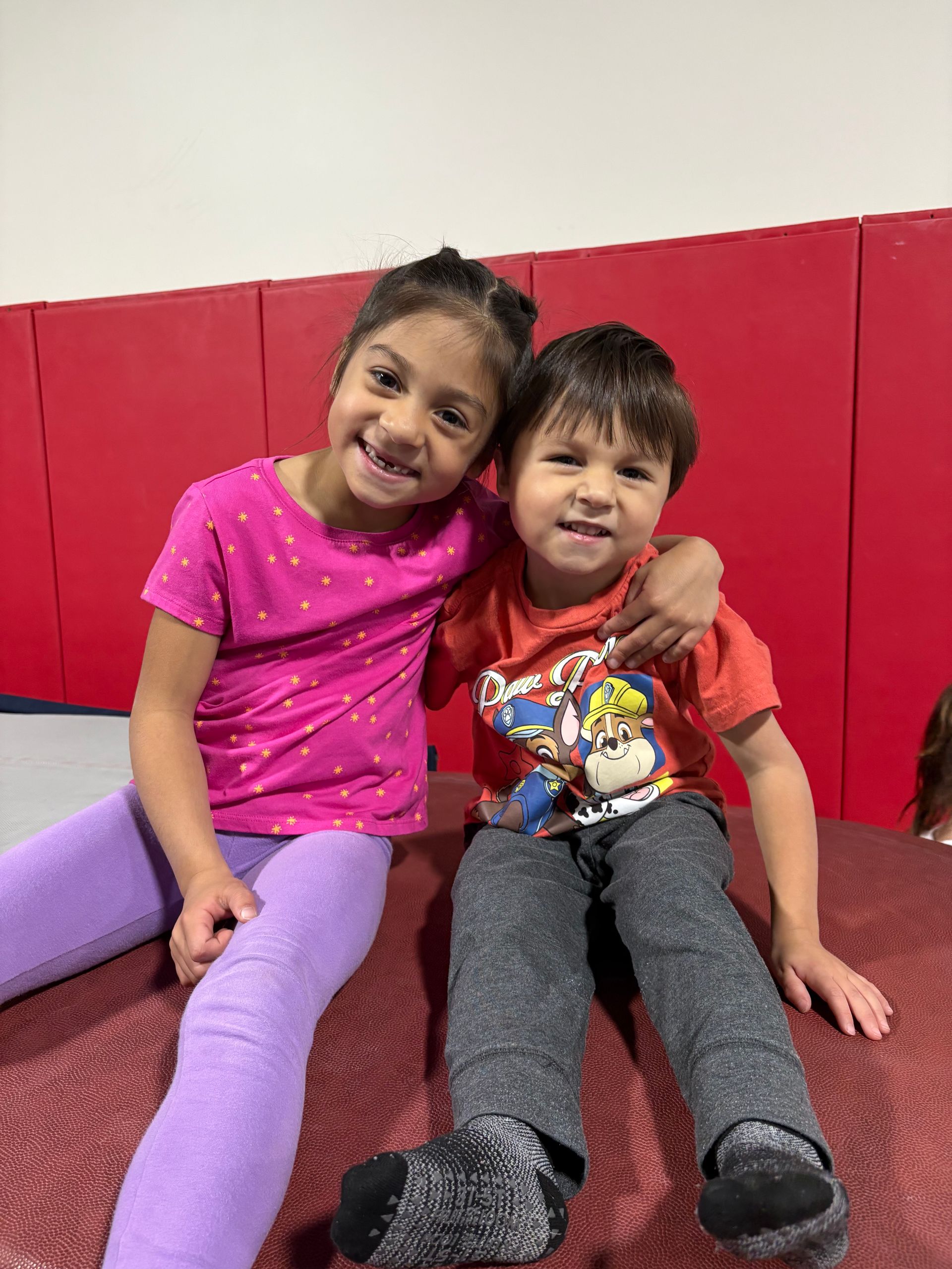 Two children sitting together on a red mat, smiling. Girl with pink shirt, purple pants; boy with orange shirt, gray pants.