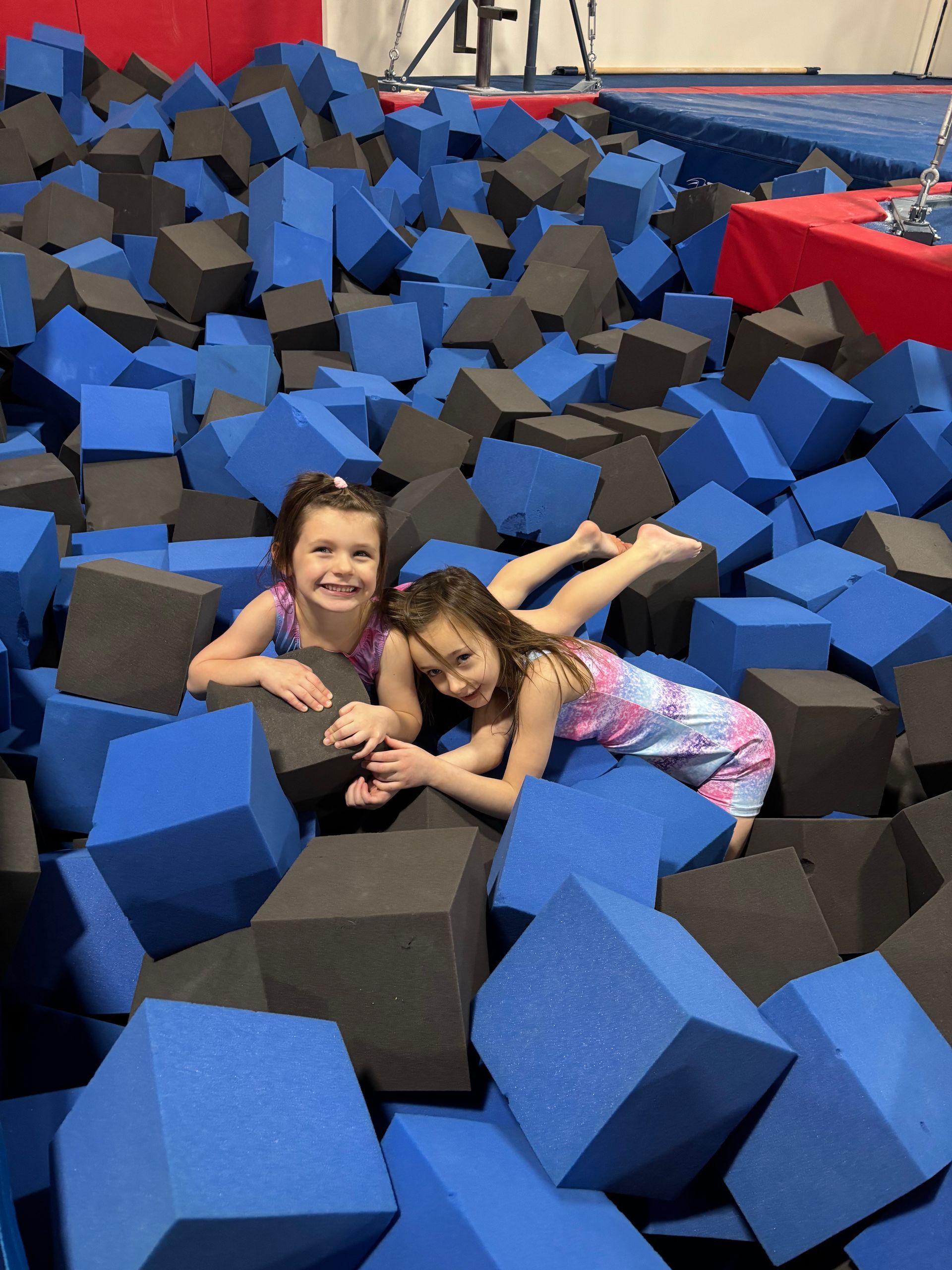 Two girls smiling in a foam pit filled with blue and black cubes at a gym.