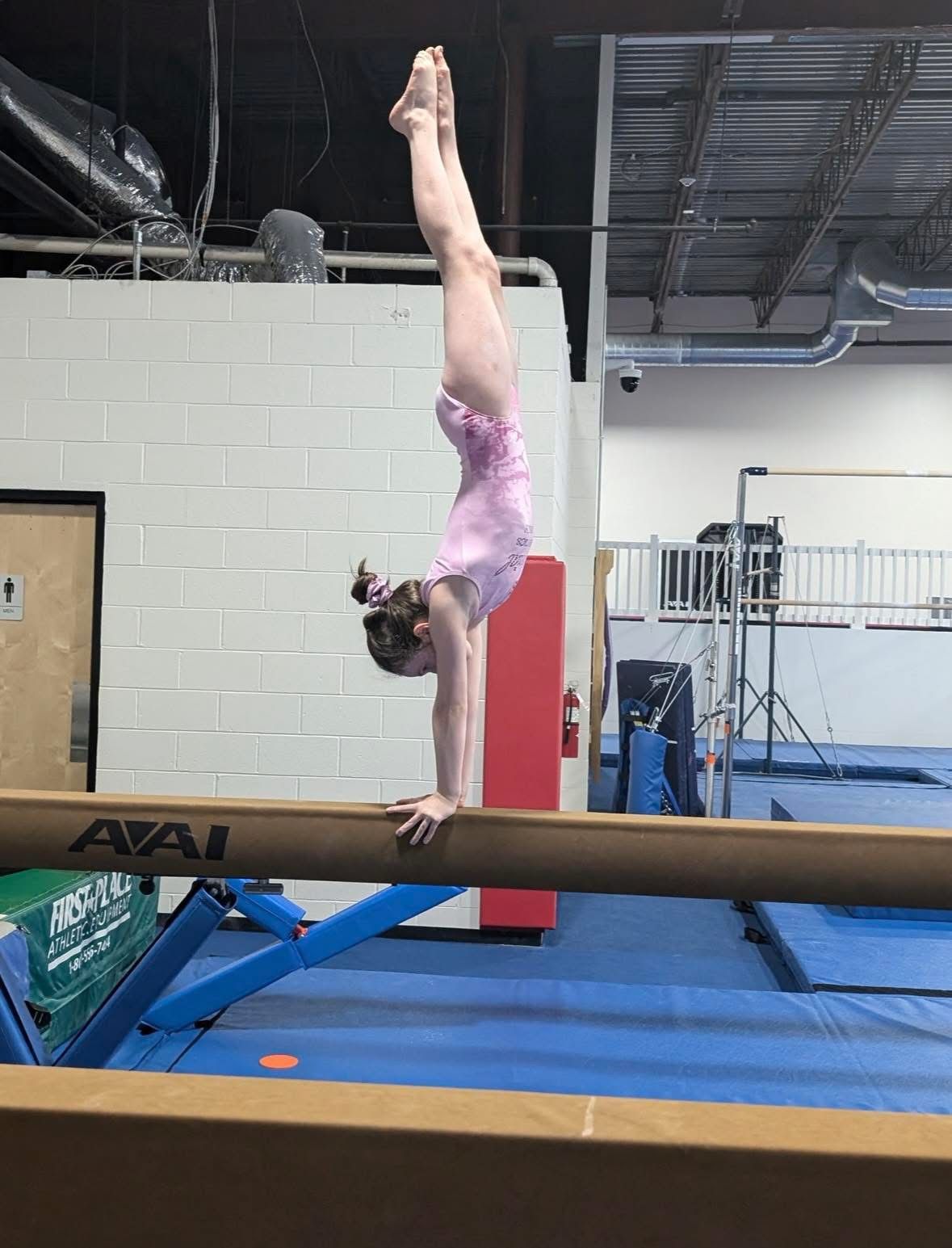 Gymnast in a pink leotard doing a handstand on a balance beam.