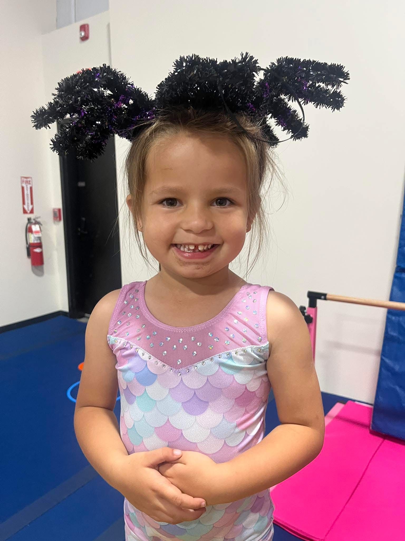 Young girl smiles, wearing a purple leotard and spider-like headpiece in a gym.