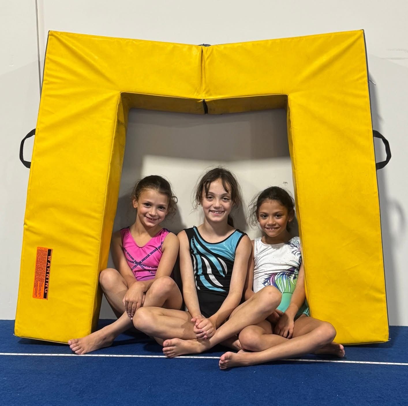 Three young gymnasts sitting inside a yellow padded archway, smiling.