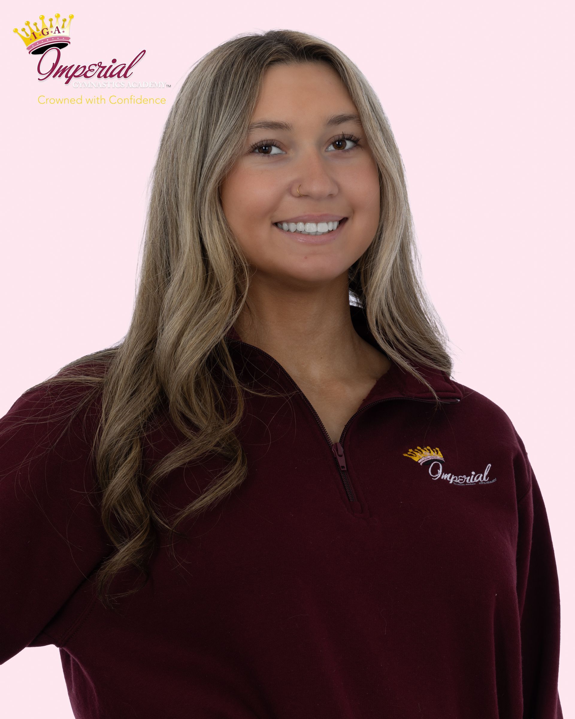 Woman with long brown hair smiling at the camera, wearing a white shirt, pink necklace, seated indoors.
