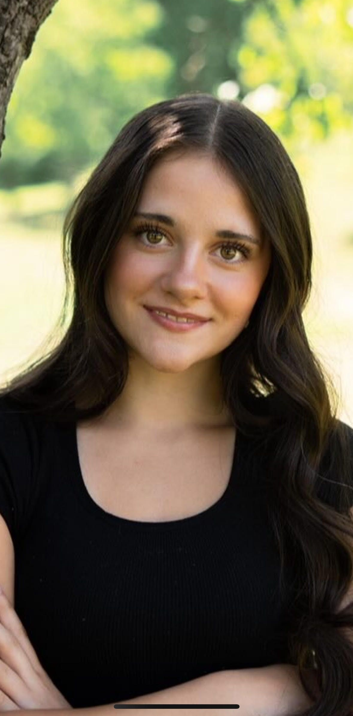 A young person with dark wavy hair smiles outdoors, wearing a black shirt.