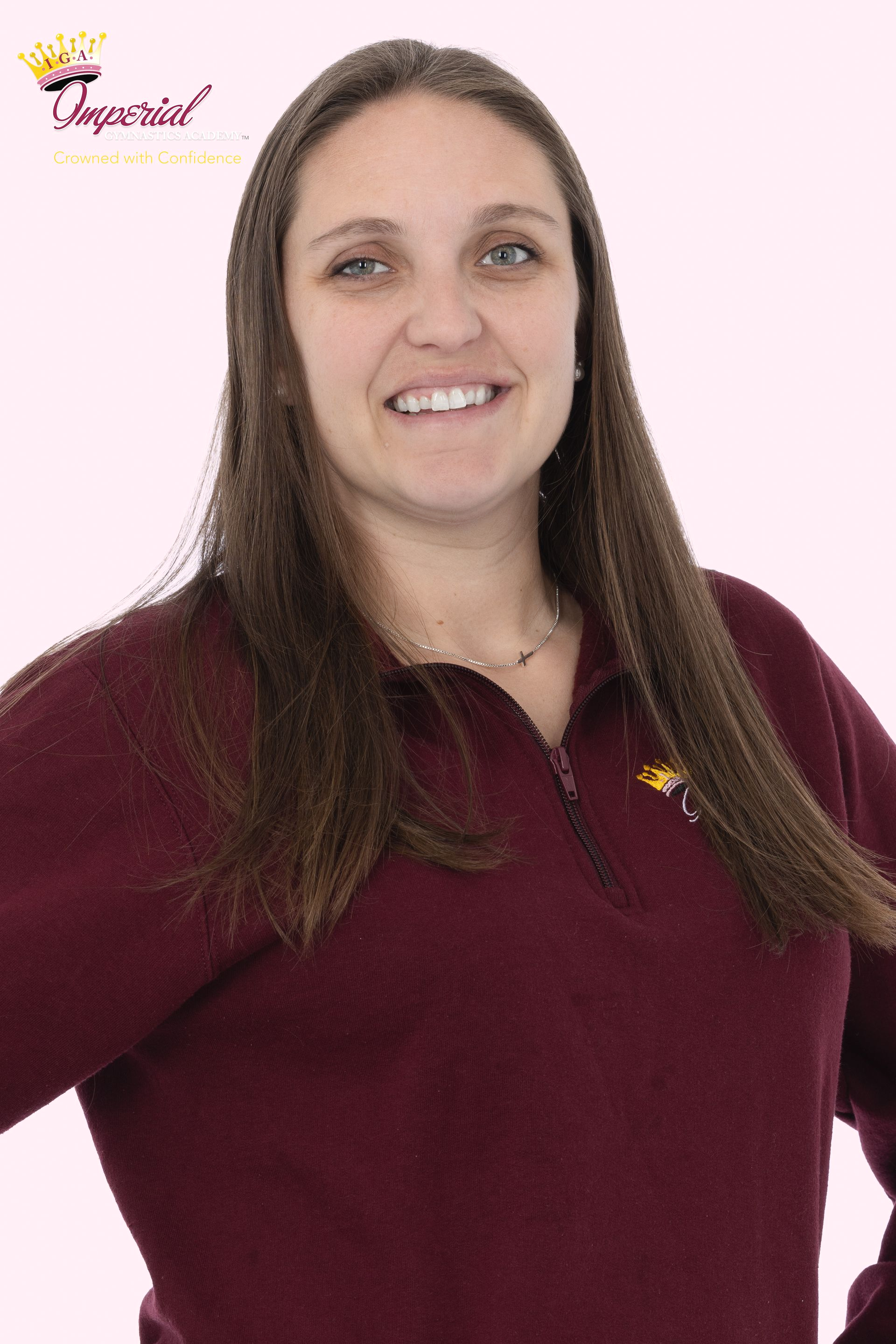Woman with brown hair smiles, wearing a pink shirt with a logo, against a light background.