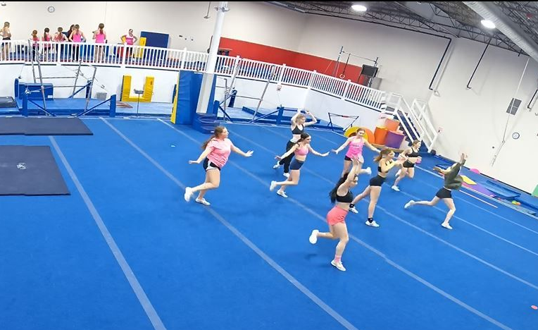 Cheerleaders in pink and black outfits running and practicing in a gym on blue mats.