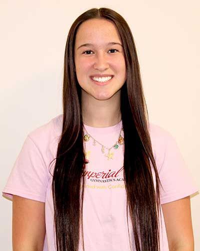 Woman with long dark hair smiles, wearing a pink t-shirt with text, necklace, against a white backdrop.
