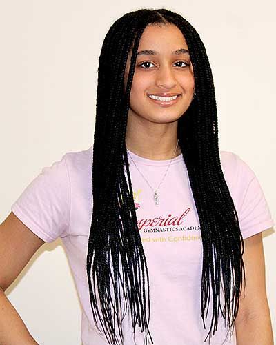 Woman with long black braids smiles, wearing a pink shirt with text, standing against a white wall.