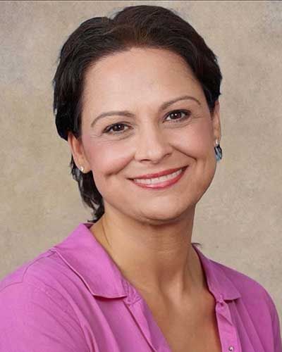 Woman with dark hair smiles, wearing a pink shirt, against a neutral background.