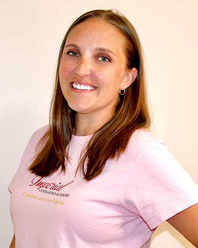 Woman with brown hair smiles, wearing a pink shirt with a logo, against a light background.