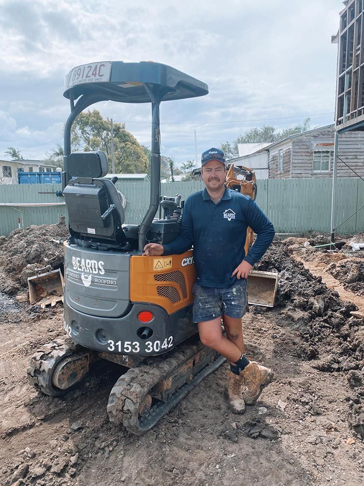 A Man is Standing Next to a Small Excavator on a Construction Site — Beards Roofing and Plumbing in Clontarf, QLD