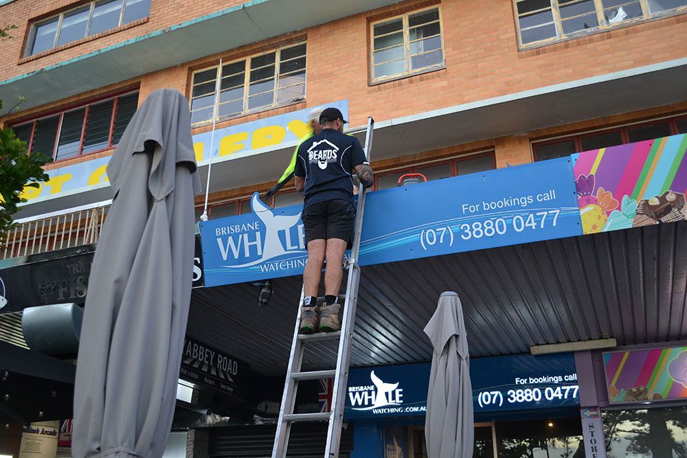 A Man is Standing on a Ladder in Front of a Building — Beards Roofing and Plumbing in Clontarf, QLD