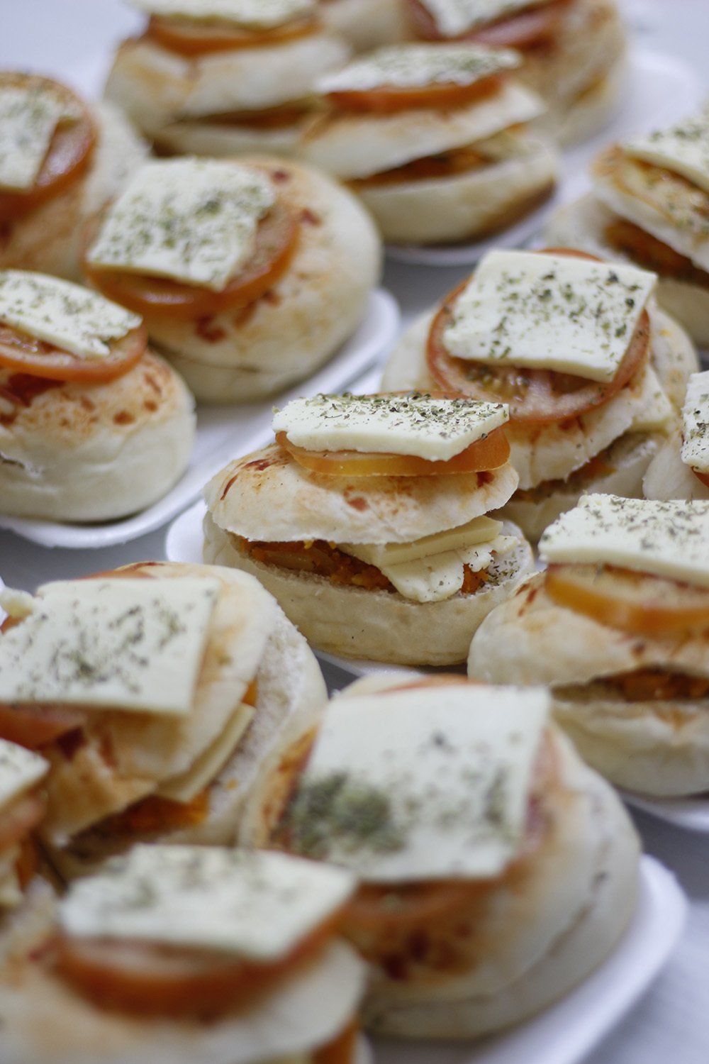 Mini sándwiches en bandejas blancas: pan redondo, rodaja de tomate, cuadrado de queso con hierbas.