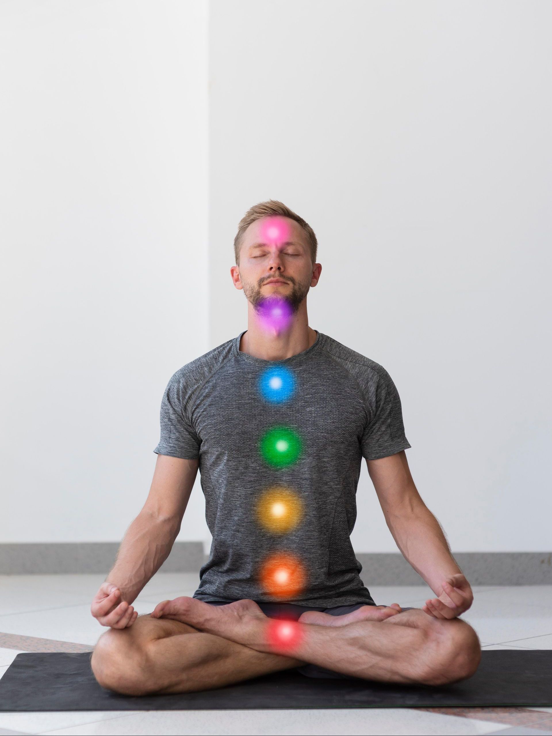 A man is sitting in a lotus position on a yoga mat with chakras surrounding his body.