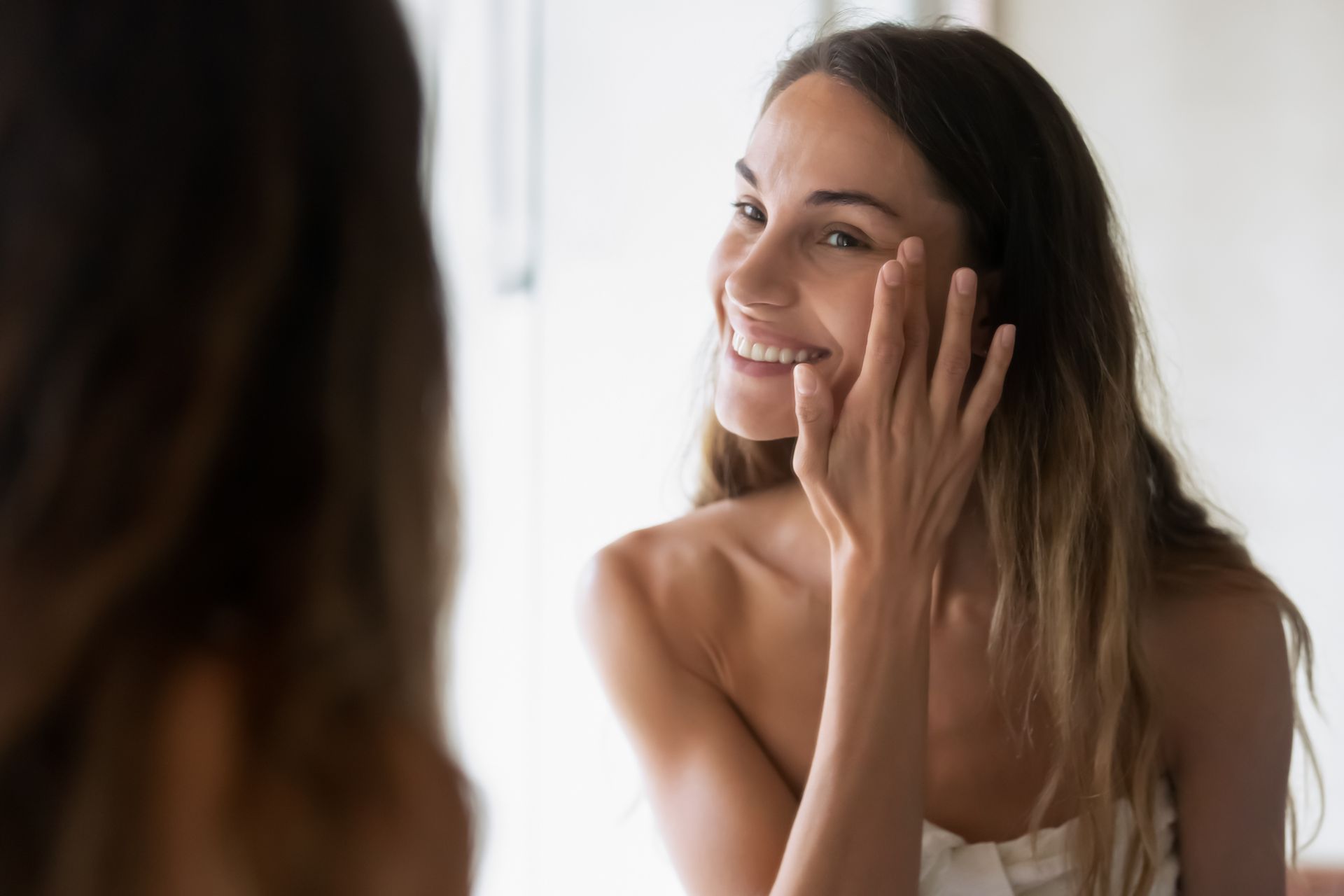 A woman is smiling while looking at her face in the mirror.
