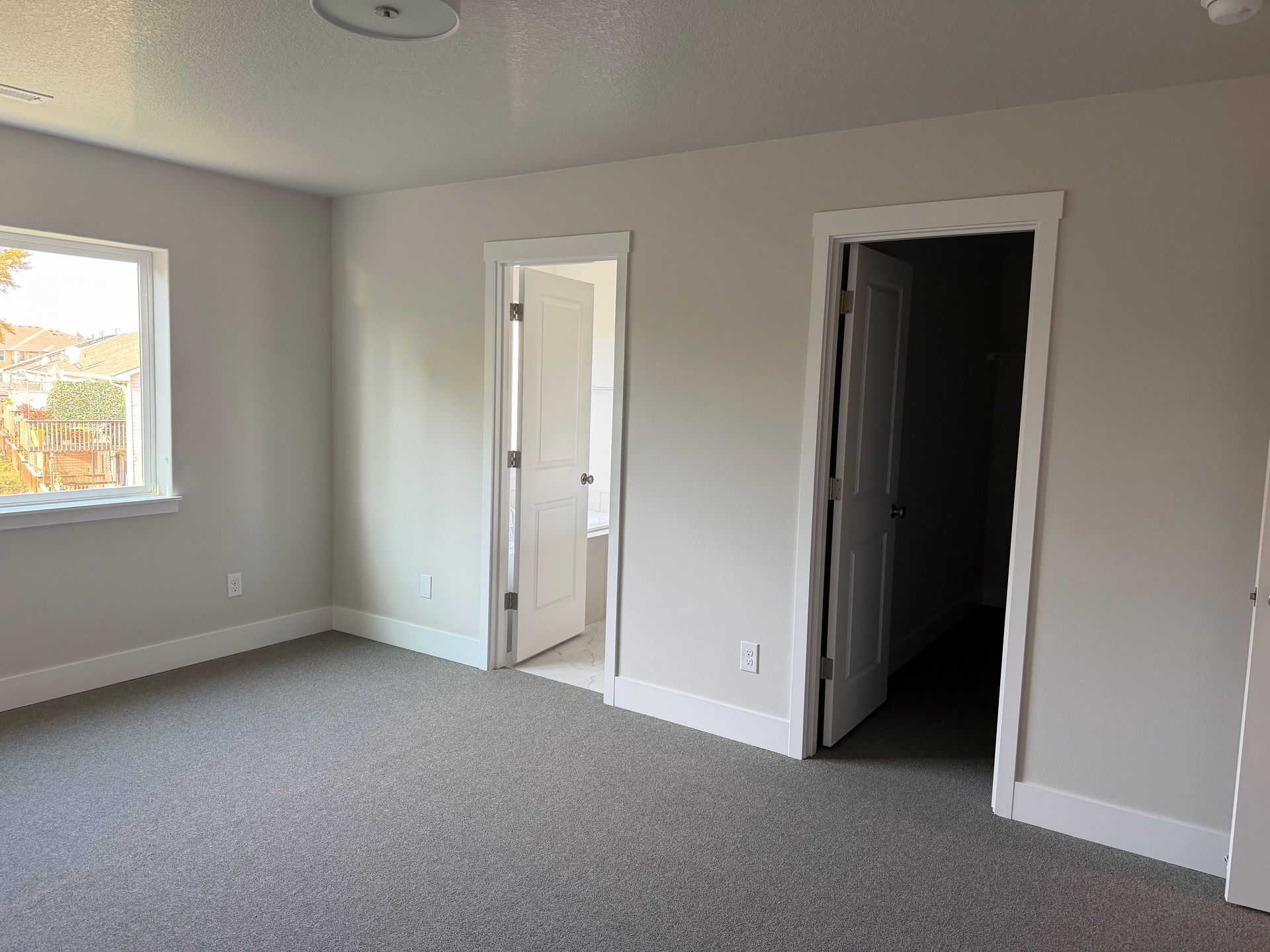 Empty bedroom with gray carpet, light gray walls, white trim, two doorways, and a window.