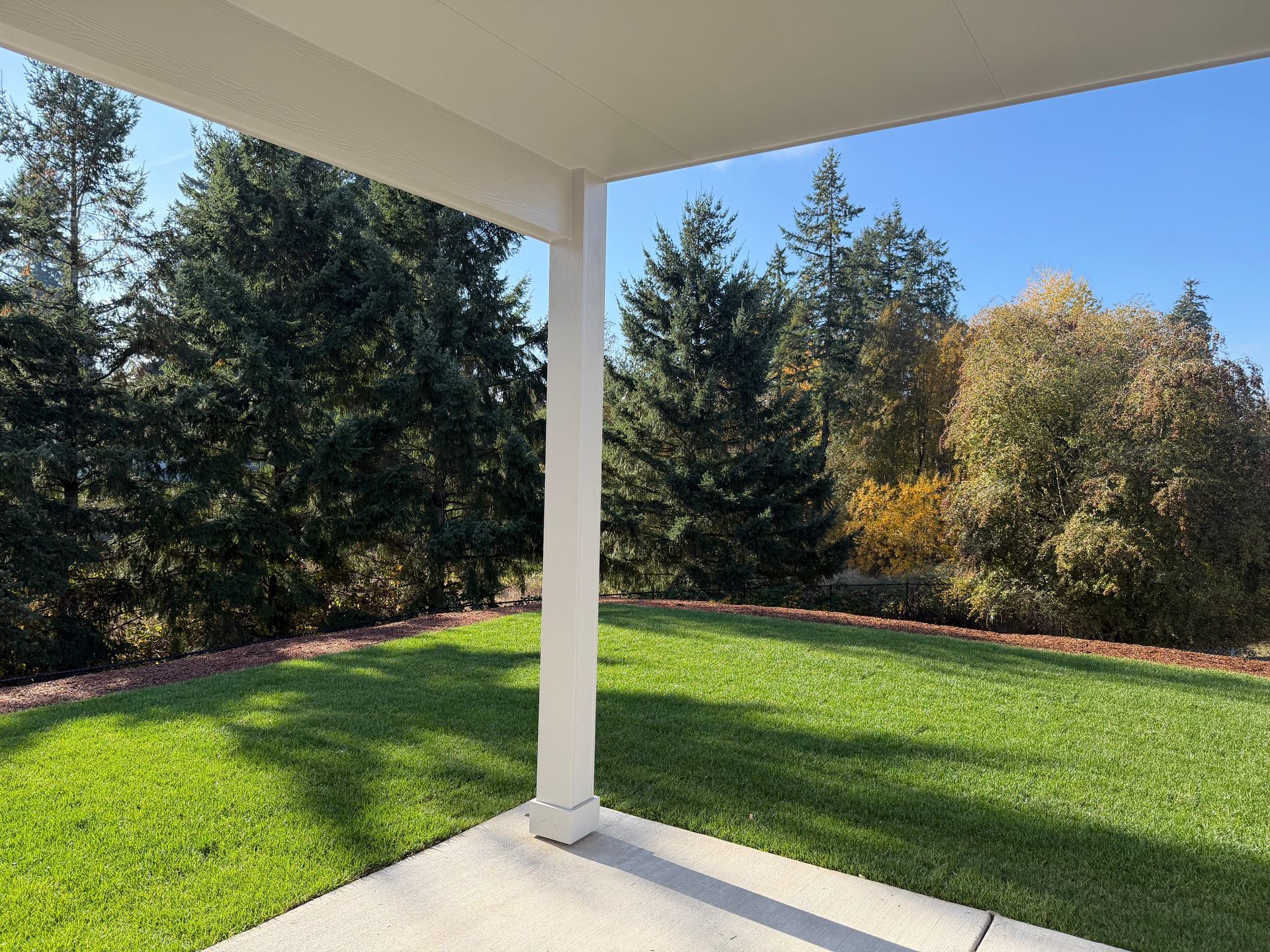 A covered patio overlooks a grassy lawn and trees under a clear blue sky.