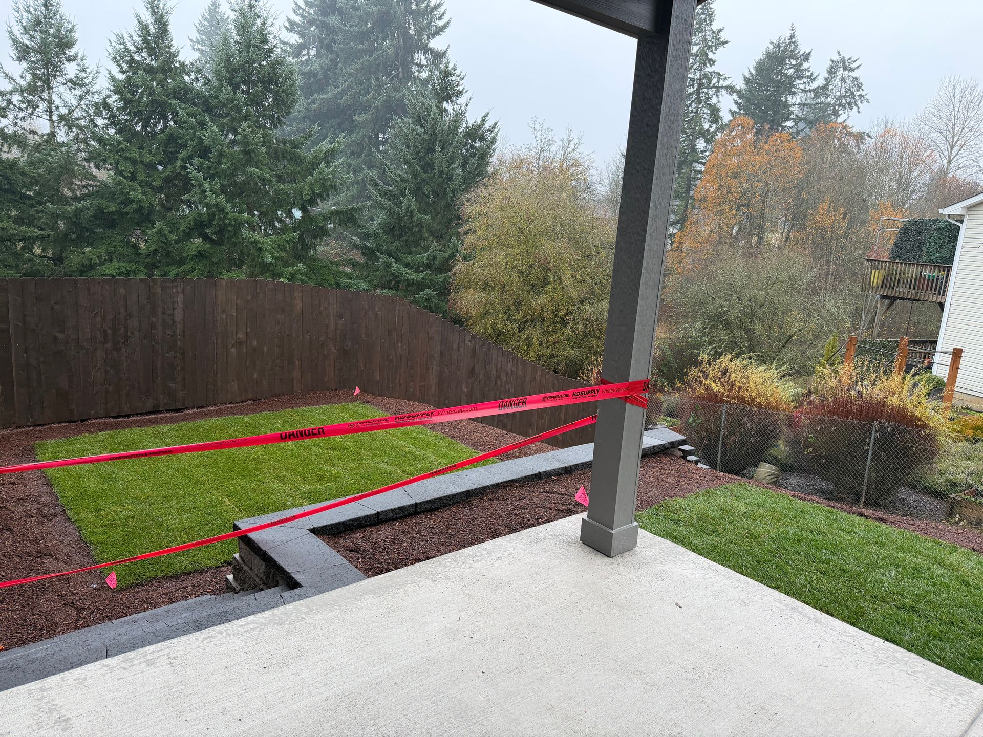 Red caution tape strung across a backyard with artificial grass, a wooden fence, and a concrete patio.