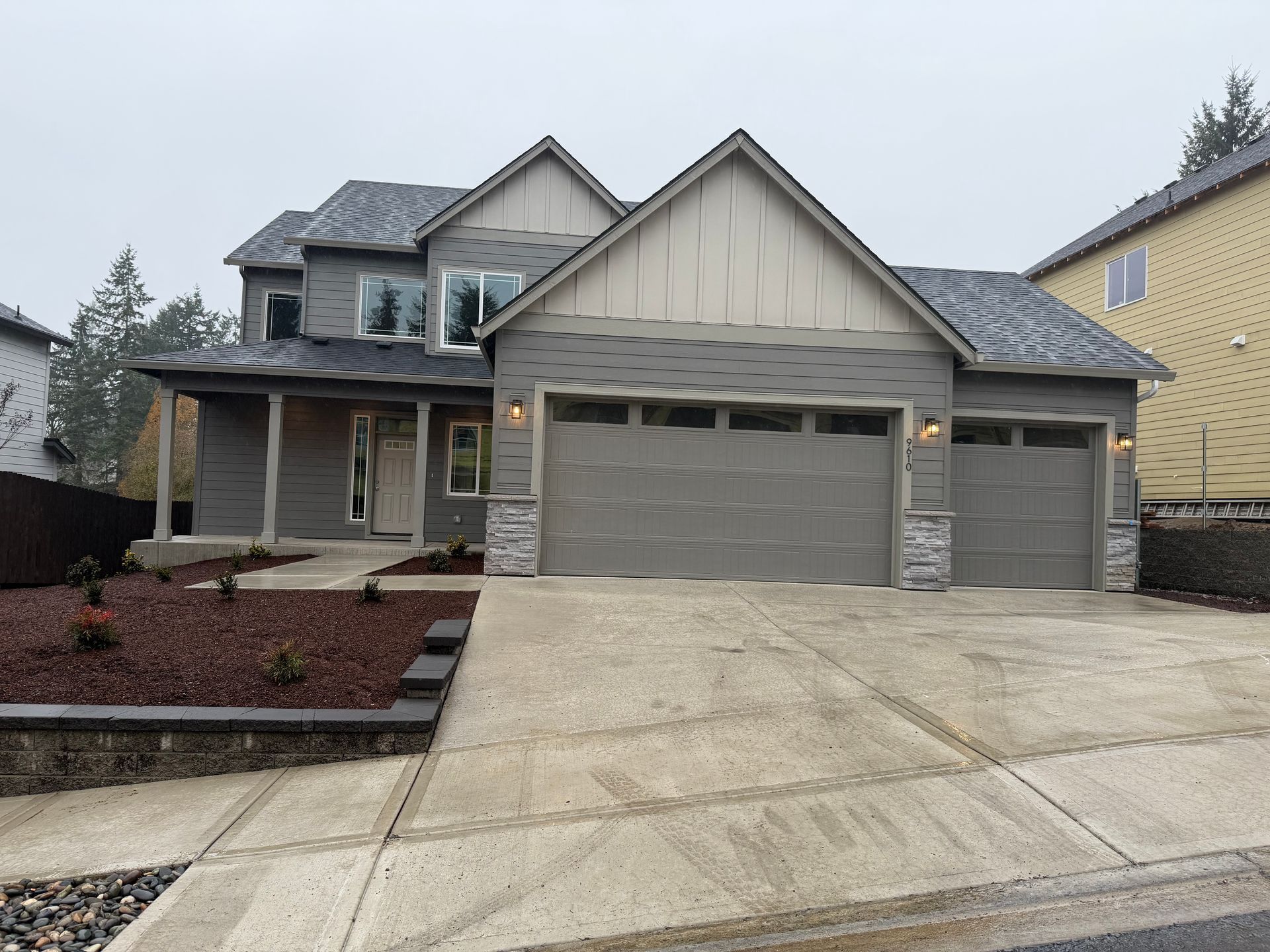 Gray two-story house with a front porch, garage, and landscaping on an overcast day.