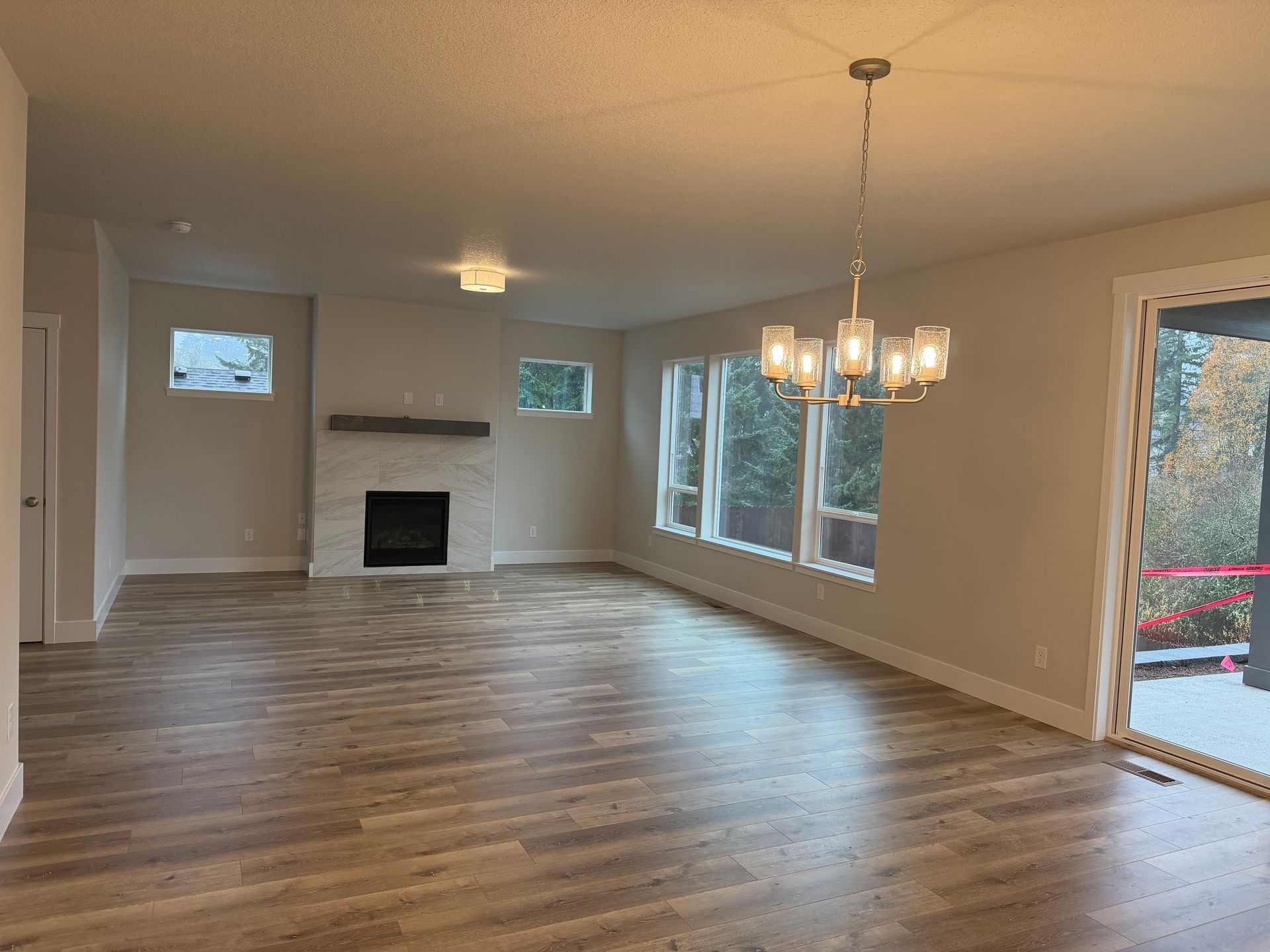 Empty living room with wood floors, fireplace, and chandelier. Large windows look out to greenery.