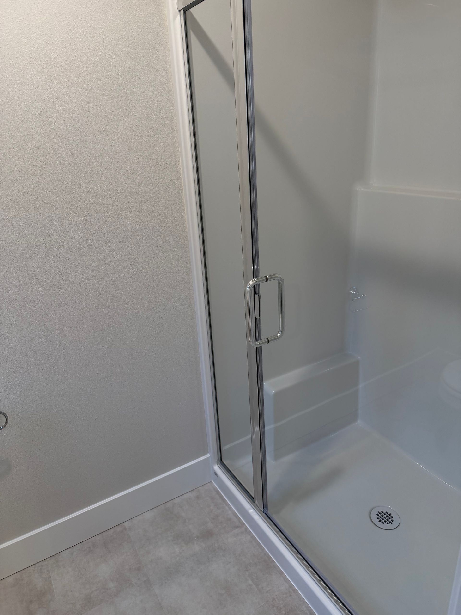 Glass shower door in a white-walled bathroom with a gray tiled floor.