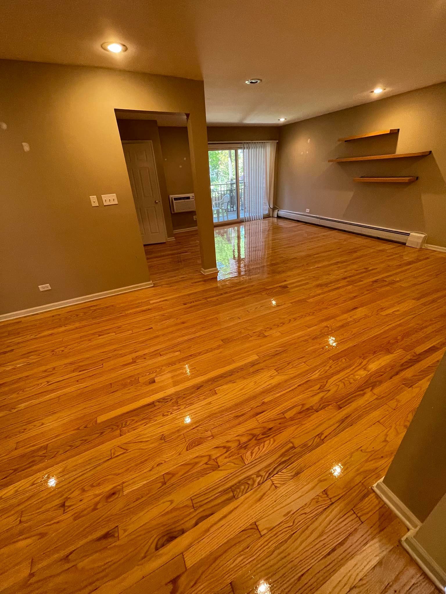 Empty room with wooden floors, neutral walls, and a sliding glass door. Two small shelves on the wall.