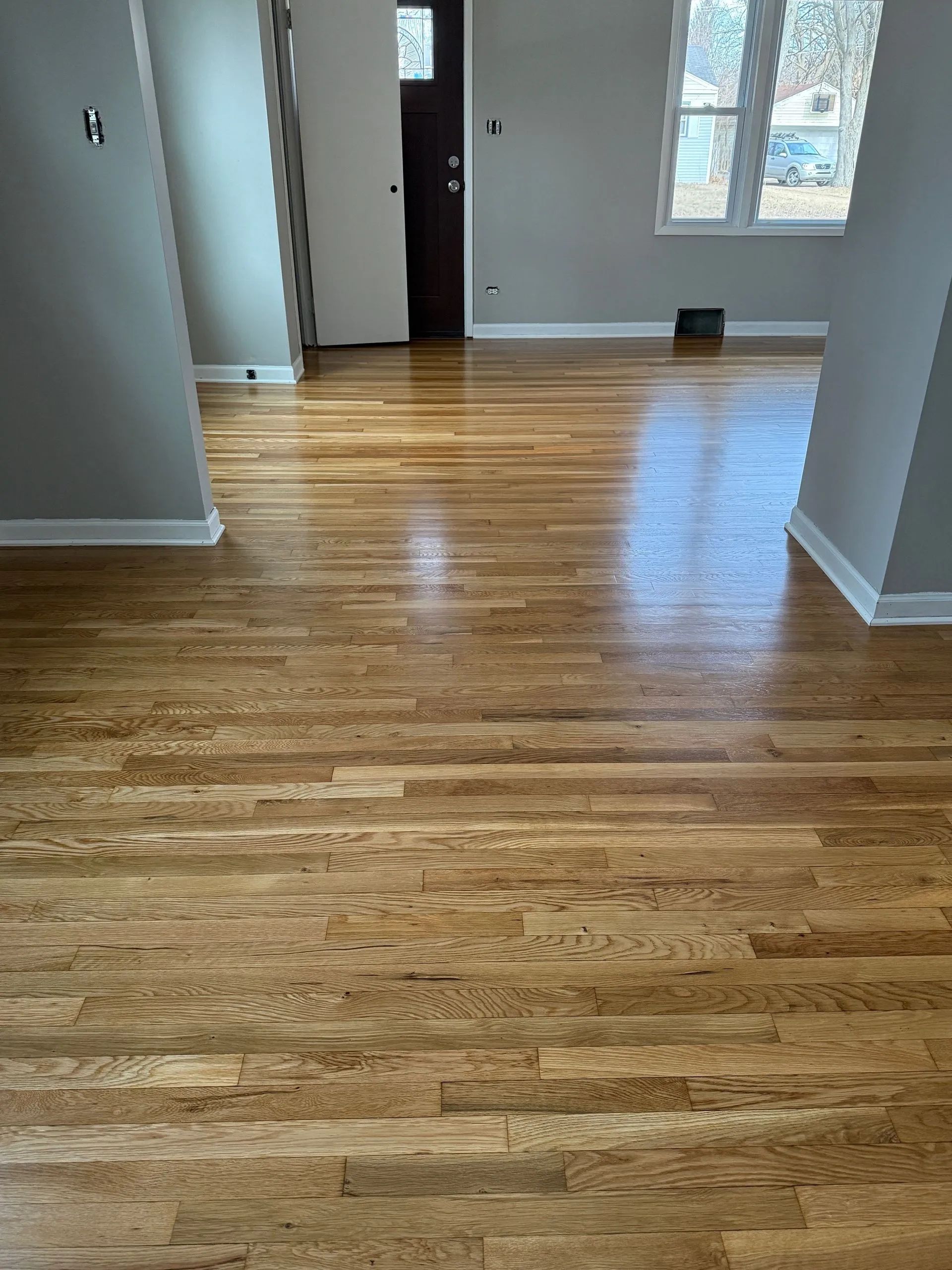 Hardwood floors in a room with gray walls and white trim, leading towards a doorway and window.